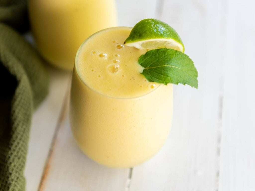 A glass of pineapple smoothie garnished with a lime wedge and a mint leaf, placed on a white wooden surface.