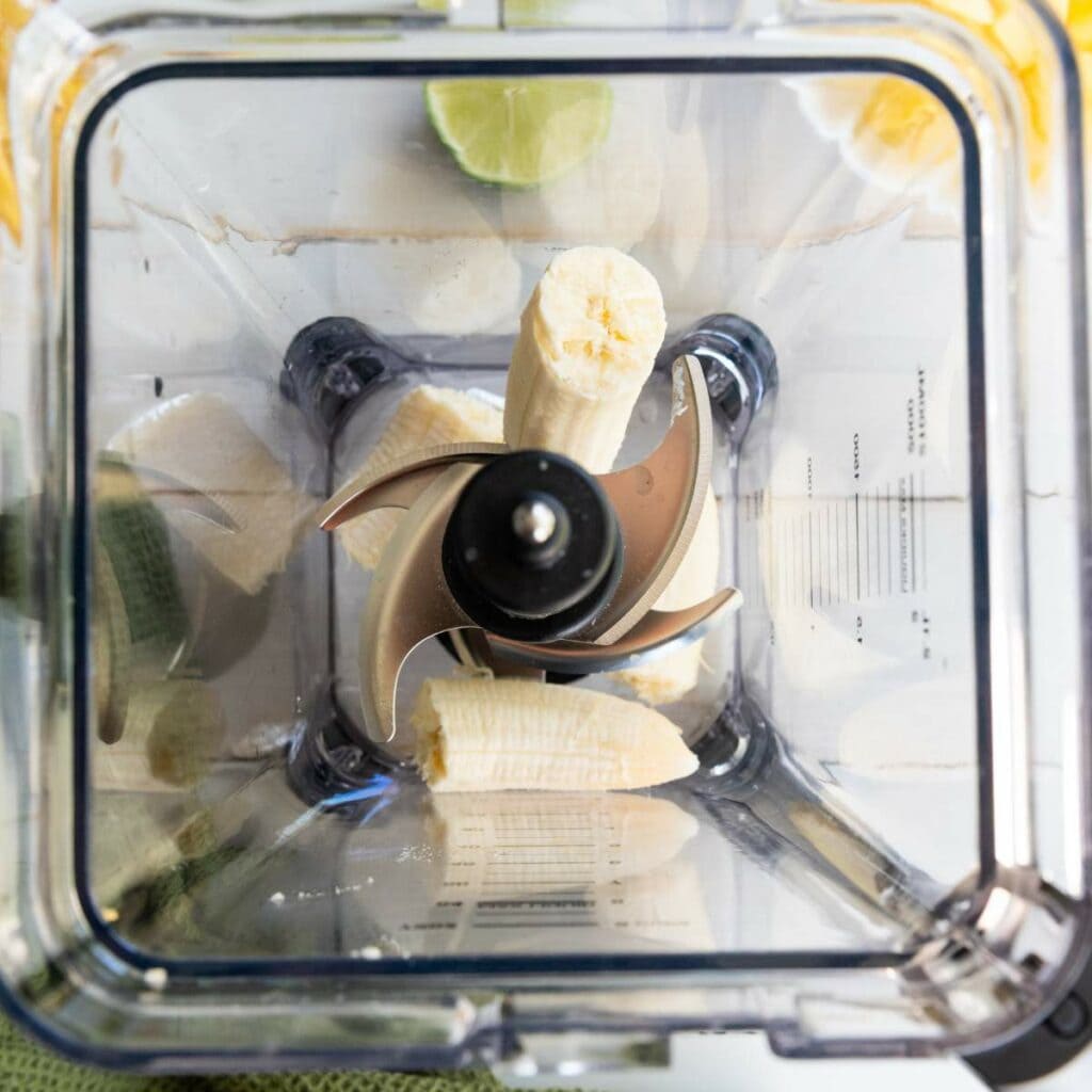 A peeled banana cut in pieces inside a blender, viewed from above, with lime halves visible around the edges.
