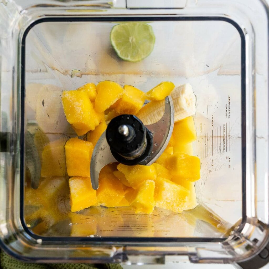 Chunks of frozen mango and a piece of banana in a blender, viewed from above, with half a lime nearby.