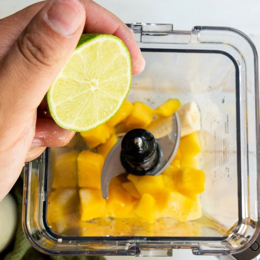A hand squeezes a lime half over a blender containing chopped mango and other ingredients.