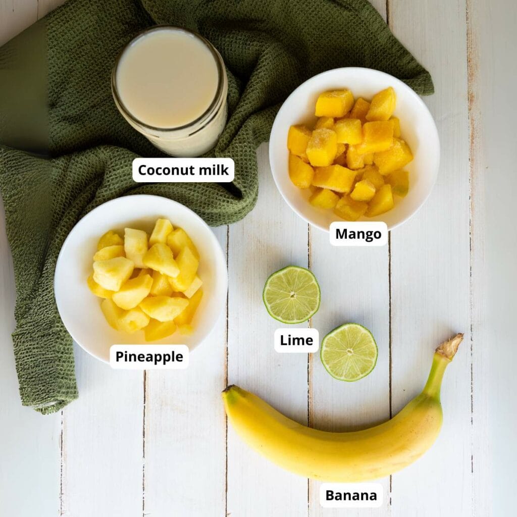 A glass of coconut milk, bowls of chopped mango and pineapple, a sliced lime, and a banana.