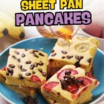 A plate of sheet pan pancakes cut into squares, topped with strawberries, bananas, and chocolate chips, with syrup being poured over them.