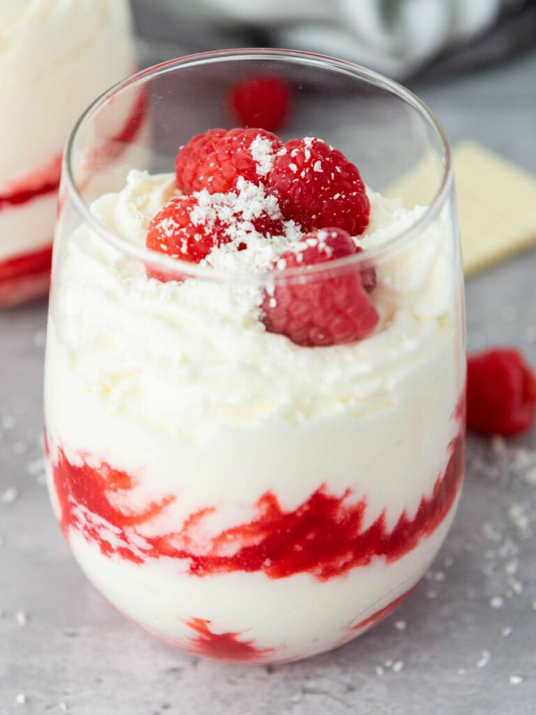 A glass filled with white chocolate mousse, topped with fresh raspberries and white chocolate shavings.