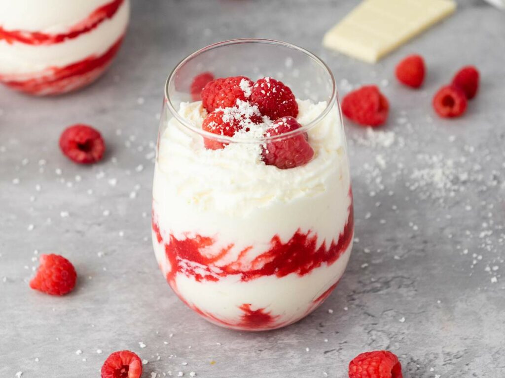 A glass filled with white chocolate mousse, topped with whole raspberries and grated white chocolate.
