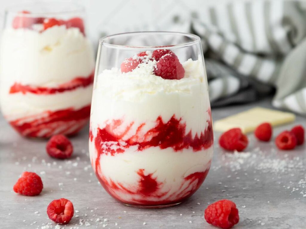 A glass filled with white chocolate moussee, topped with raspberries and shredded coconut, with raspberries scattered on the surface.