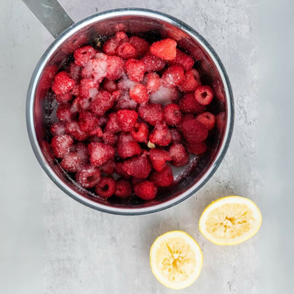 A saucepan filled with raspberries and sugar sits on a gray surface next to two squeezed lemon halves.