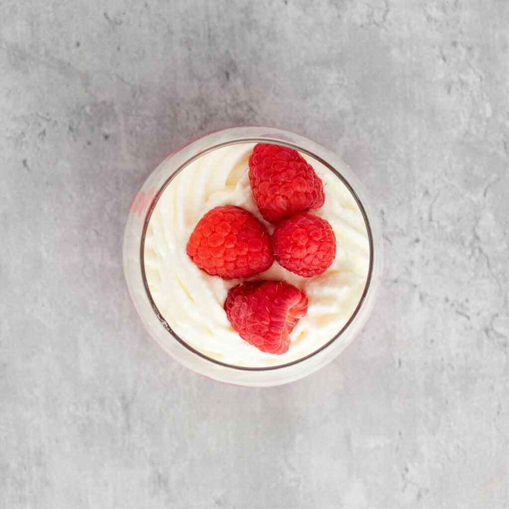 A glass cup filled with swirled whipped cream, topped with four raspberries, on a light gray textured surface.