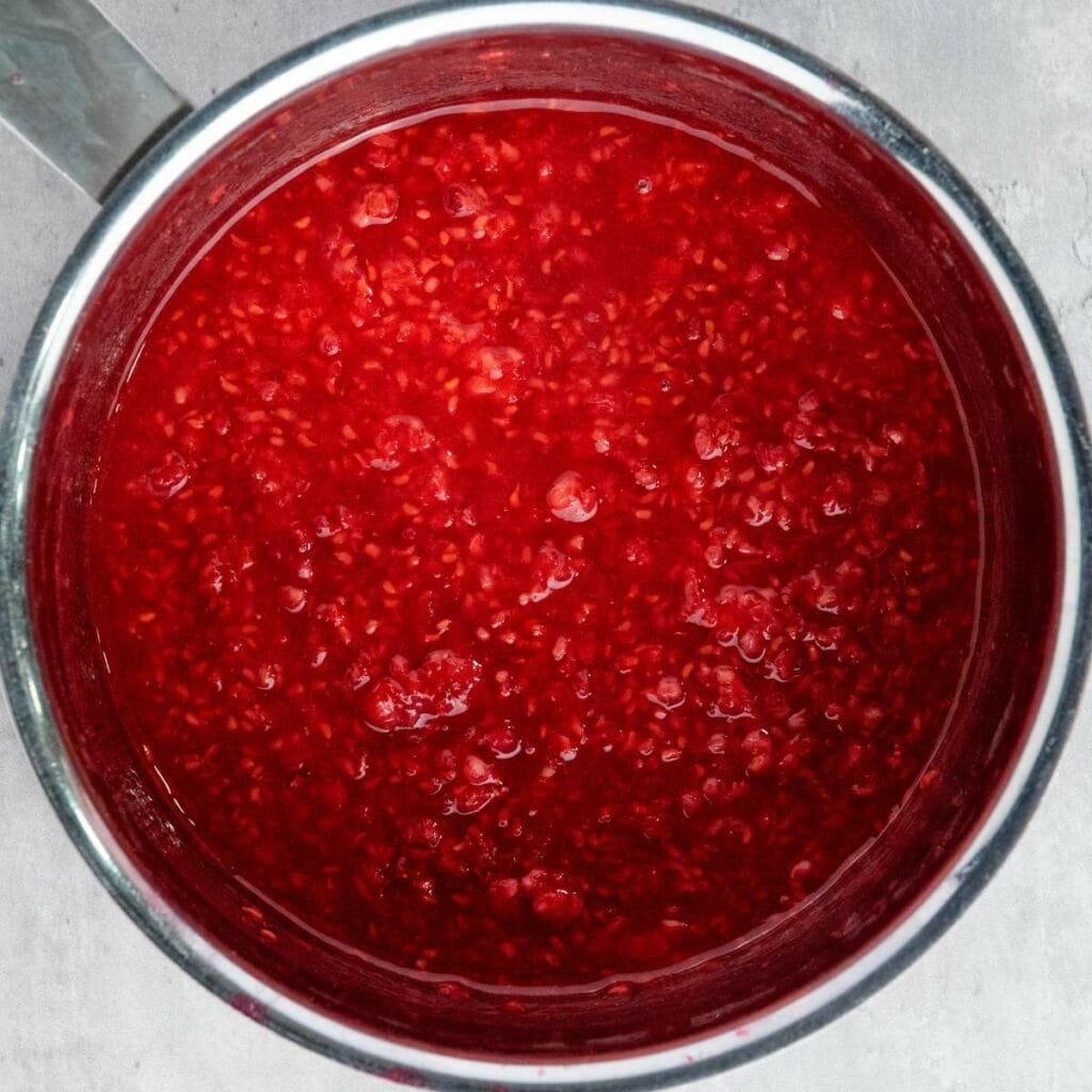 A stainless steel pot filled with thick, red raspberry sauce or compote with visible seeds and fruit pieces.