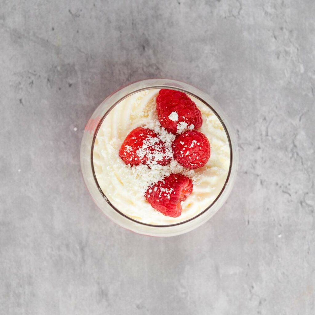 A glass cup filled with whipped dessert topped with three raspberries and a dusting of powdered sugar, set on a gray textured surface.