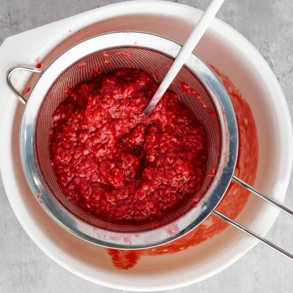 A metal strainer with mashed raspberries and a spoon rests over a white bowl, with raspberry juice collecting below.