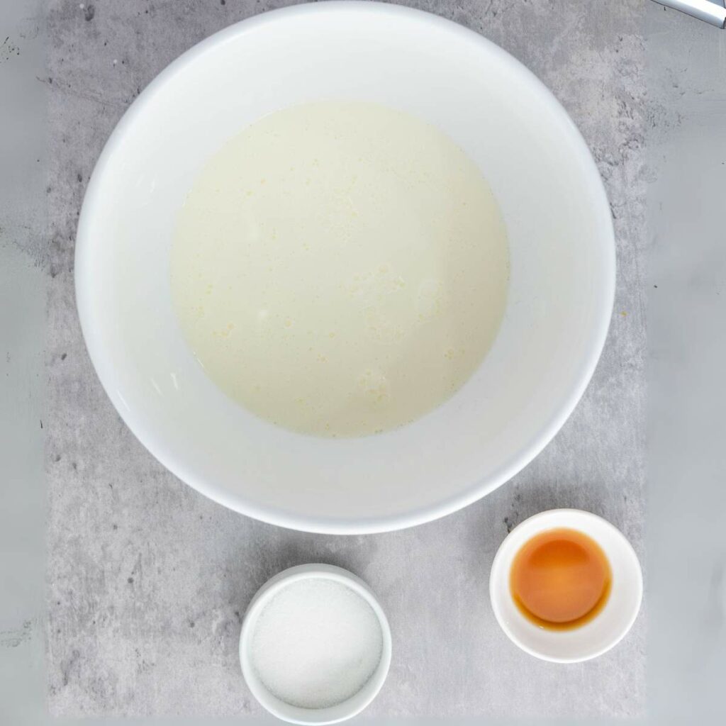 A white bowl with a creamy liquid, a small bowl of white sugar, and a small bowl of amber liquid on a gray surface.