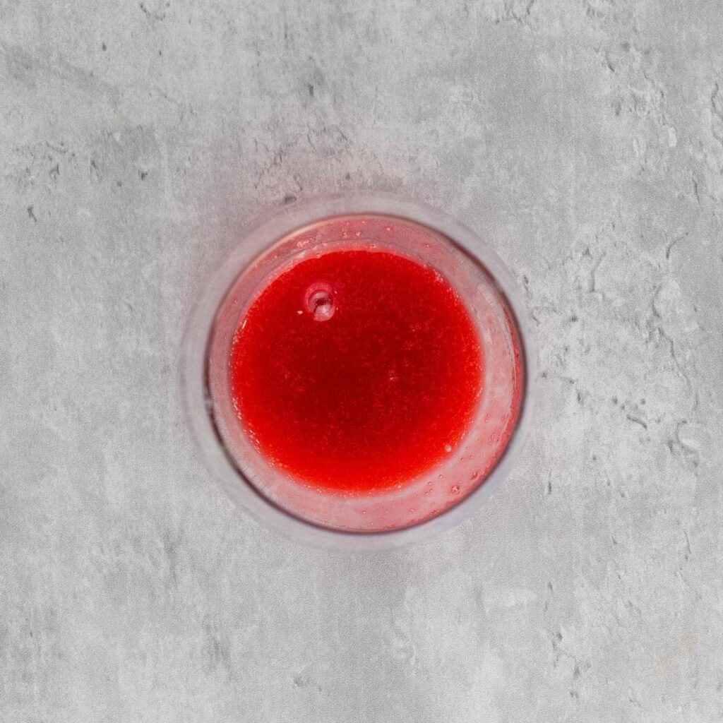 A glass filled with bright red liquid, viewed from above, placed on a light gray textured surface.