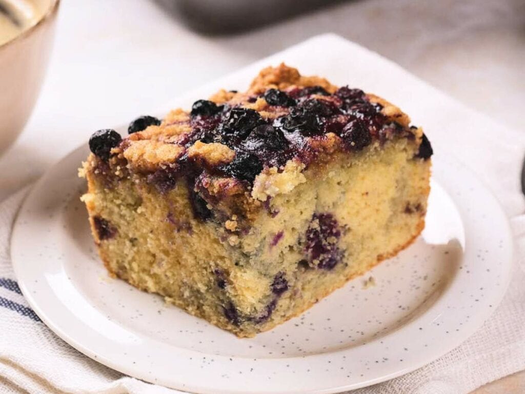 A slice of blueberry crumb cake with a golden crust and visible blueberries, served on a white plate.