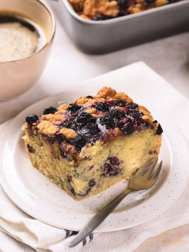 A square slice of blueberry crumb cake on a white plate with a fork, set on a napkin, with a coffee cup and baking tray in the background.