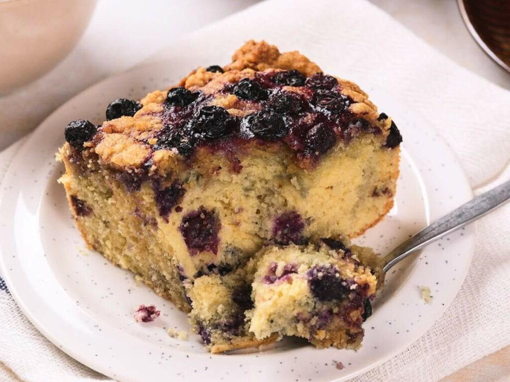 A slice of blueberry coffee cake with a crumbly topping sits on a white plate with a fork and a piece cut off.