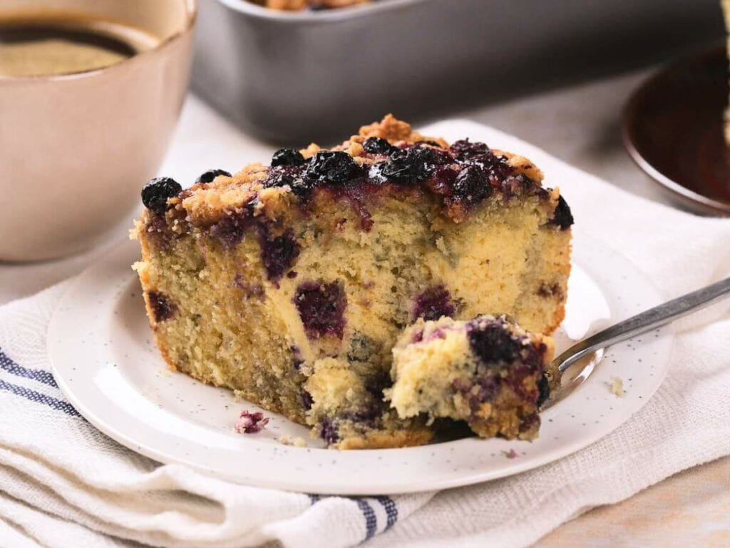 A slice of blueberry coffee cake on a white plate with a fork, next to a cup of coffee.