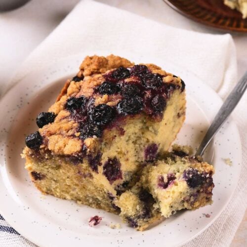A slice of blueberry coffee cake with a crumb topping is served on a white plate with a fork on a napkin.