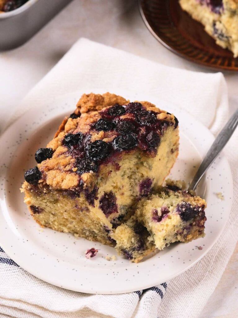 A slice of blueberry coffee cake with a crumb topping is served on a white plate with a fork on a napkin.