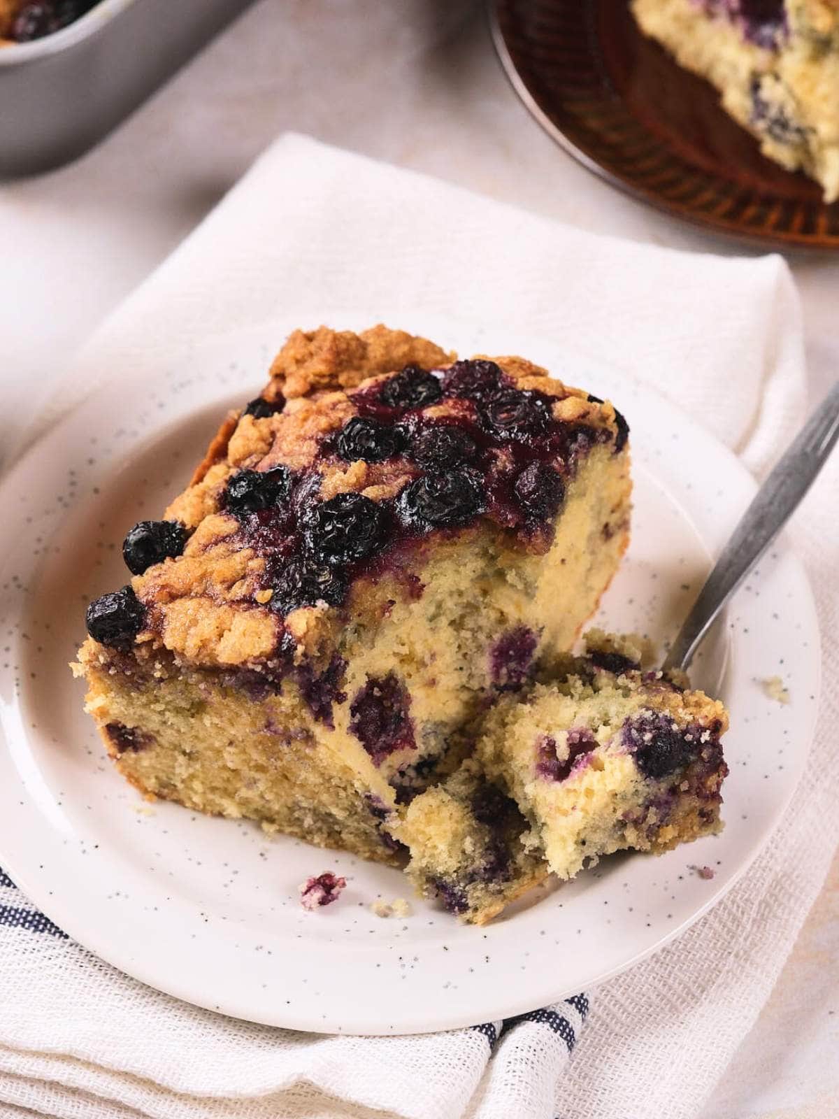 A slice of blueberry coffee cake with a crumb topping is served on a white plate with a fork on a napkin.