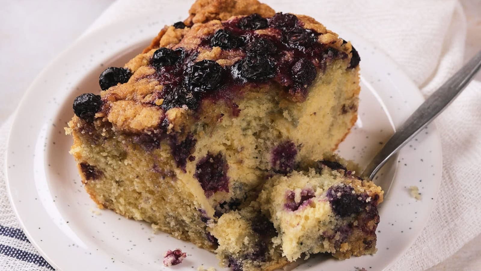 A slice of blueberry coffee cake on a white plate with a fork, showing a crumbly topping and blueberry pieces throughout the cake.