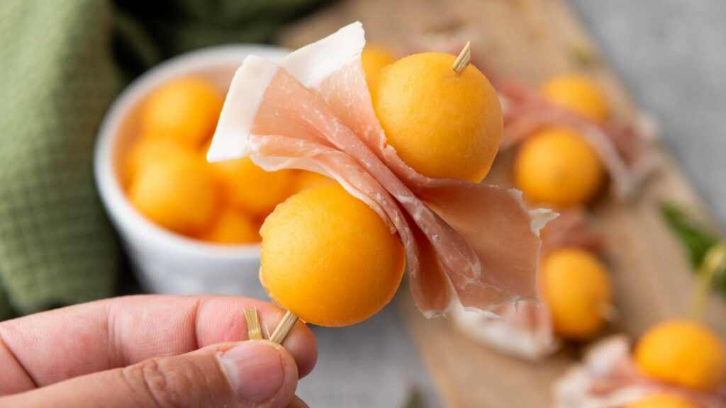 A hand holds a skewer with two cantaloupe prosciutto and a folded slice of prosciutto.