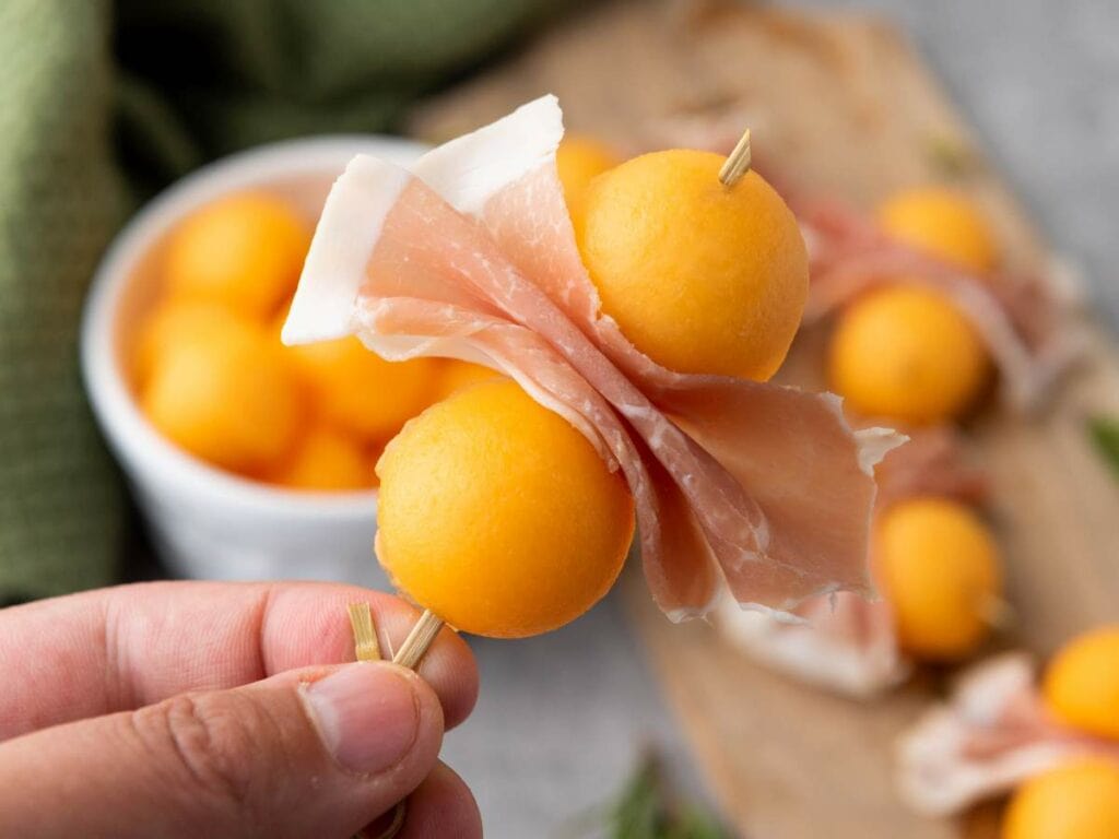 A hand holds a skewer with two cantaloupe prosciutto balls.