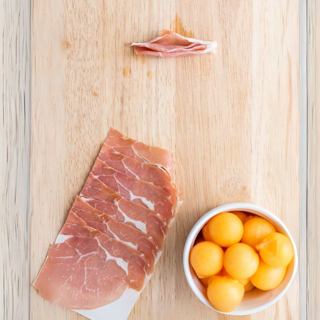 Slices of prosciutto, a small bowl of cantaloupe balls, and a single rolled piece of prosciutto.