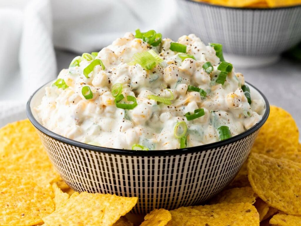 A bowl of creamy corn dip topped with chopped green onions, surrounded by yellow tortilla chips.