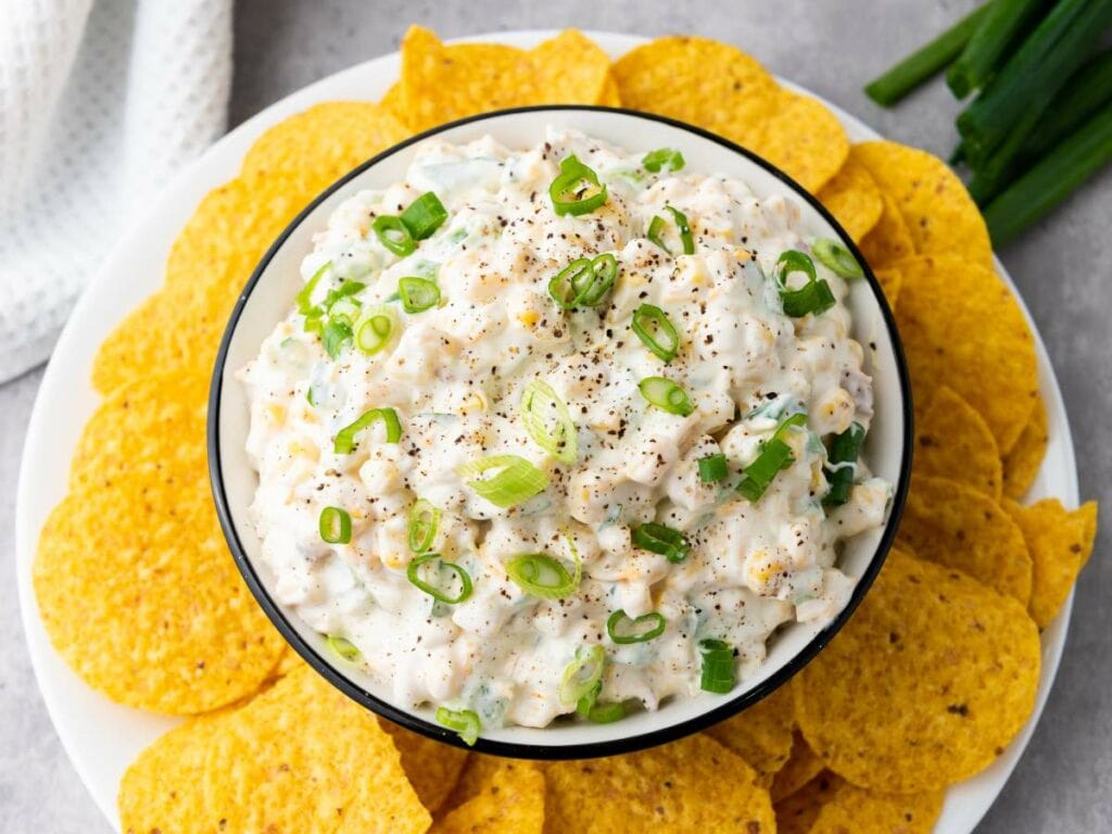 A creamy corn dip topped with chopped green onions and black pepper, surrounded by round yellow tortilla chips.