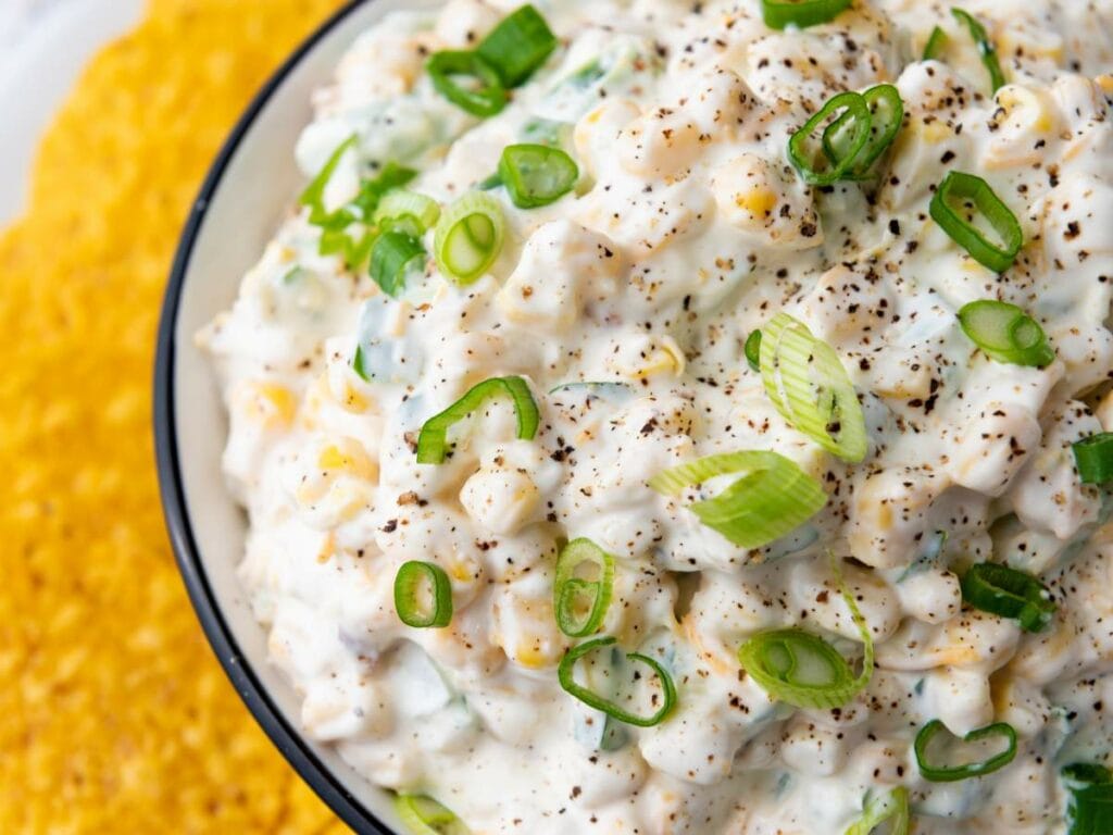A close-up of a creamy corn dip topped with sliced green onions and cracked black pepper.