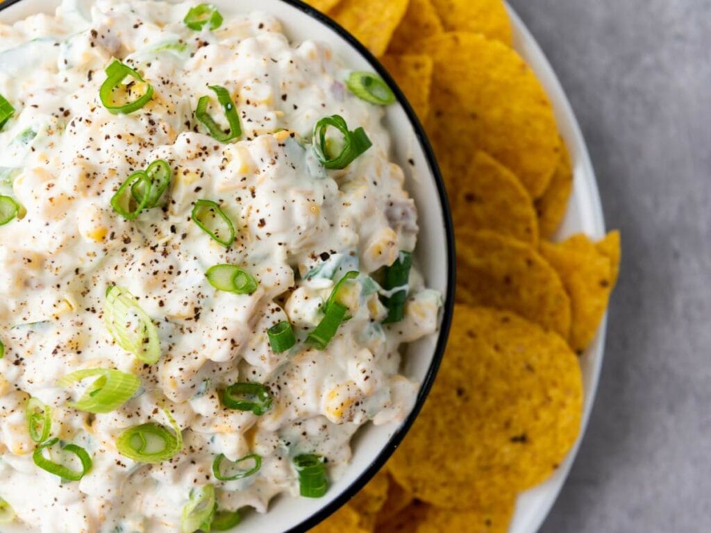 A bowl of creamy corn dip topped with chopped green onions and black pepper.