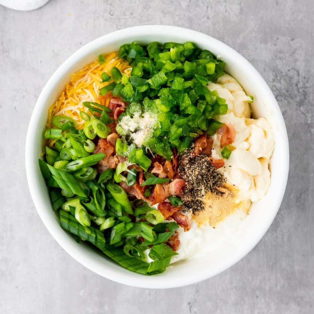 A white bowl filled with shredded cheese, chopped green onions, diced green peppers, bacon, mayonnaise, and seasoning.