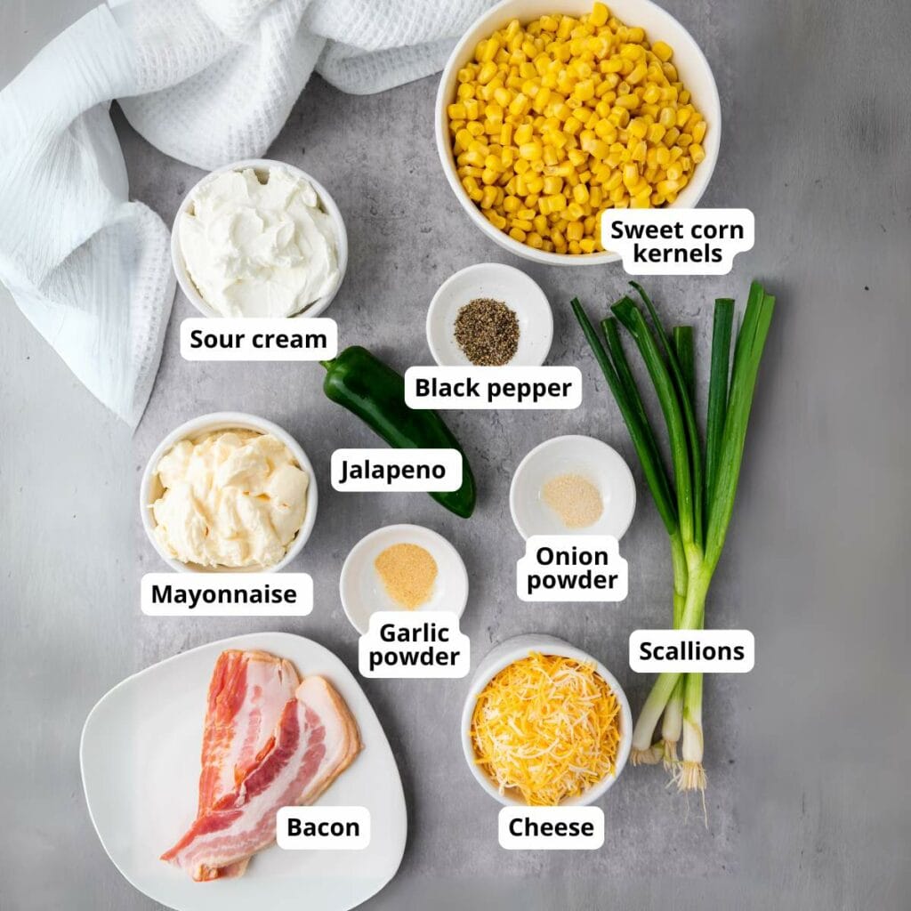 Various labeled ingredients for a corn dip recipe, including sweet corn kernels, scallions, cheese, bacon, mayonnaise, sour cream, jalapeño, black pepper, garlic powder, and onion powder.