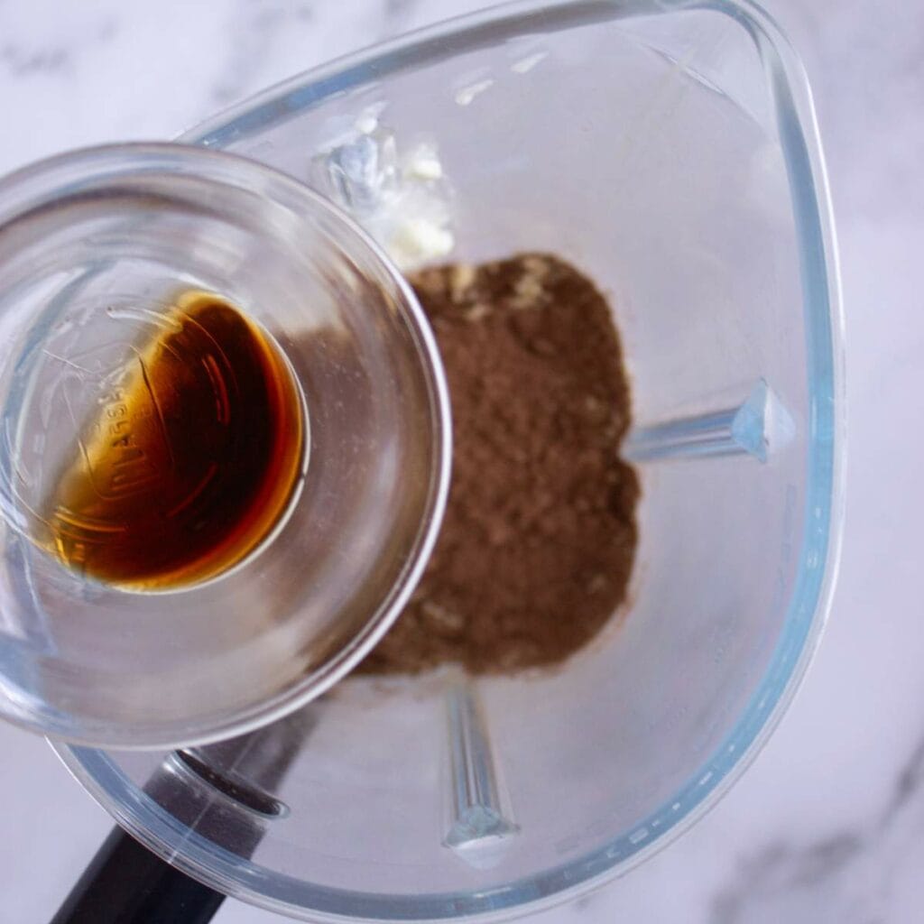 A glass blender contains cocoa powder and a small glass cup of vanilla extract.