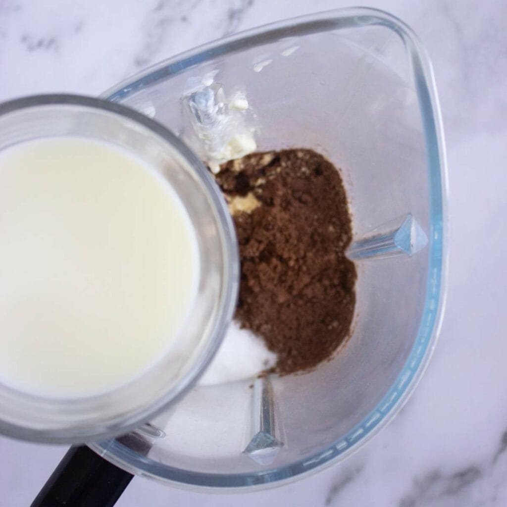 Top view of a blender containing milk, cocoa powder, sugar, and milk.
