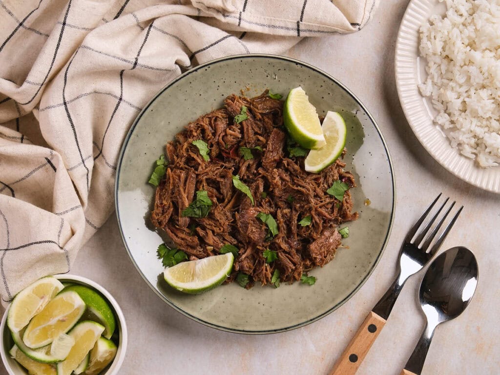 A plate of barbacoa garnished with cilantro and lime wedges, next to a bowl of lime slices.