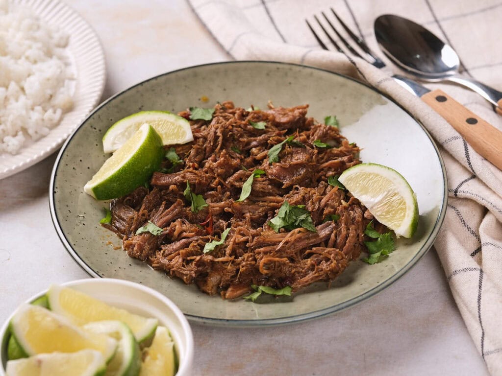 A plate of barbacoa garnished with cilantro and lime wedges, with a fork, spoon, and a bowl of lime slices nearby.