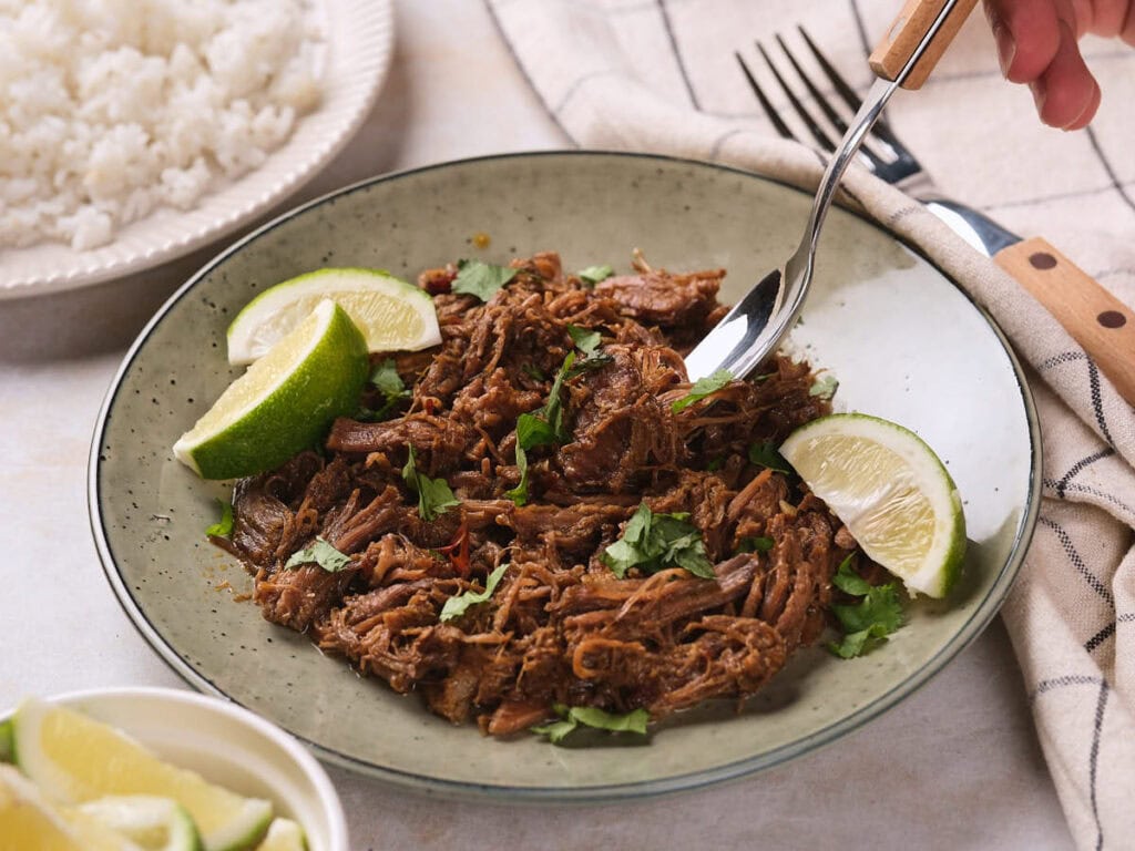 Barbacoa garnished with cilantro and lime wedges, with a fork and knife beside it.