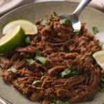 A bowl of barbacoa garnished with chopped cilantro and lime wedges, with a spoon lifting a portion from the plate.