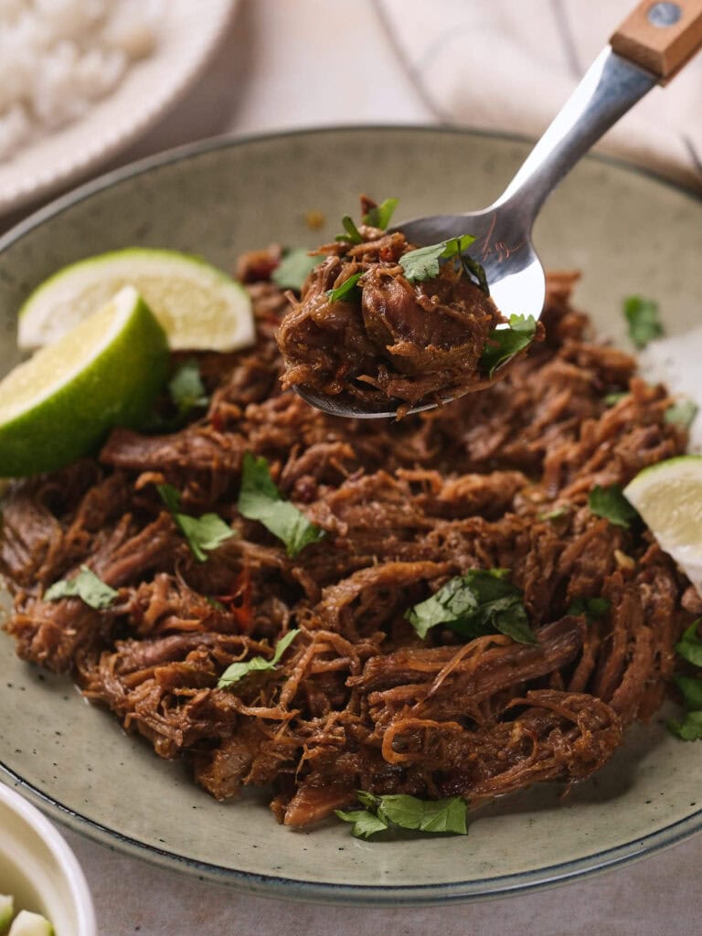 A bowl of barbacoa garnished with chopped cilantro and lime wedges, with a spoon lifting a portion from the plate.