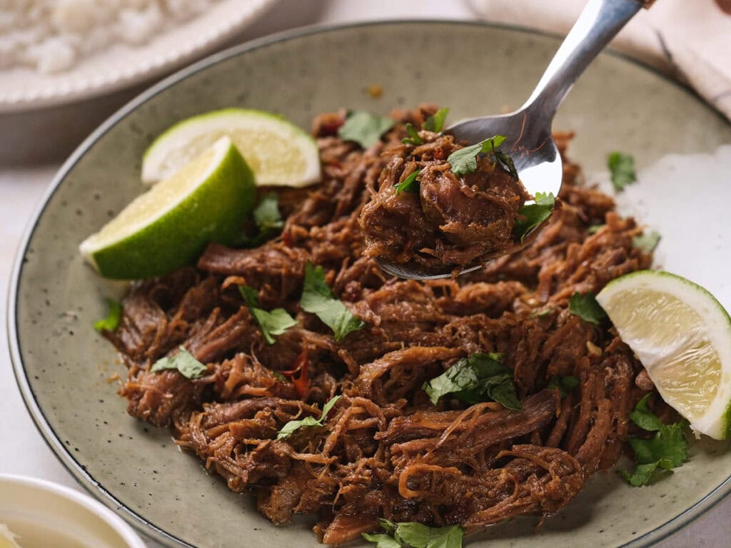 Barbacoa garnished with chopped cilantro and lime wedges, with a spoon lifting a portion.