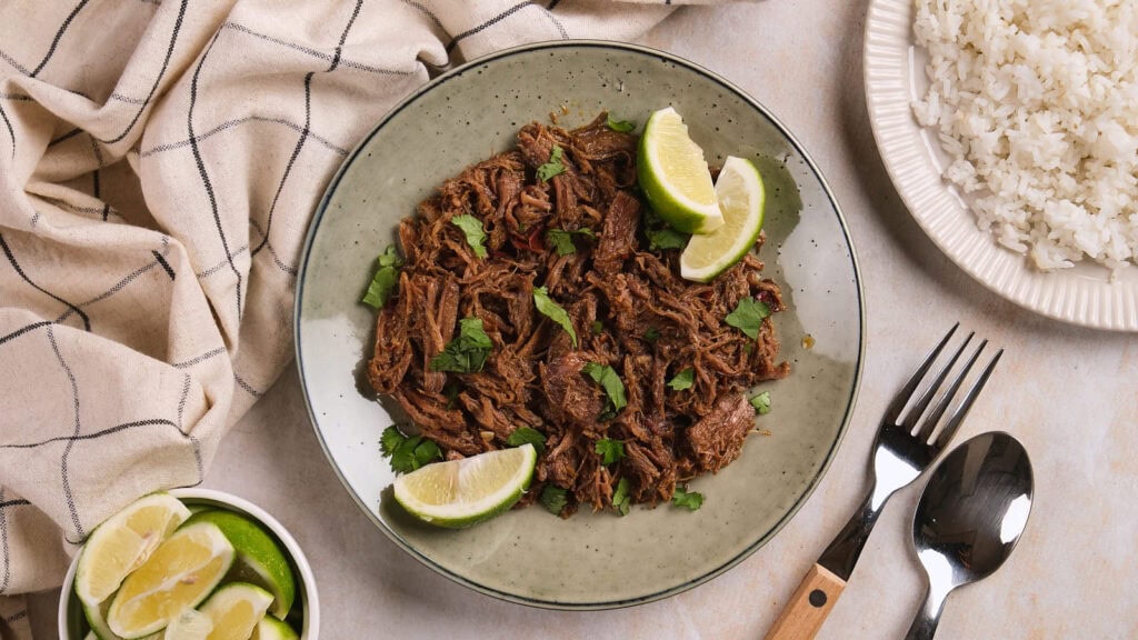 A plate of barbacoa garnished with cilantro and lime wedges, served next to a bowl of lime slices.