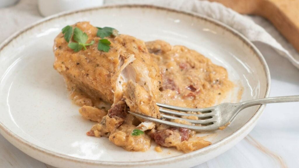 A piece of chicken breast topped with creamy sauce and herbs is served on a white plate with a fork cutting into it.