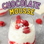 A glass of white chocolate mousse with raspberry swirls, topped with fresh raspberries and white chocolate shavings, sits on a gray surface.