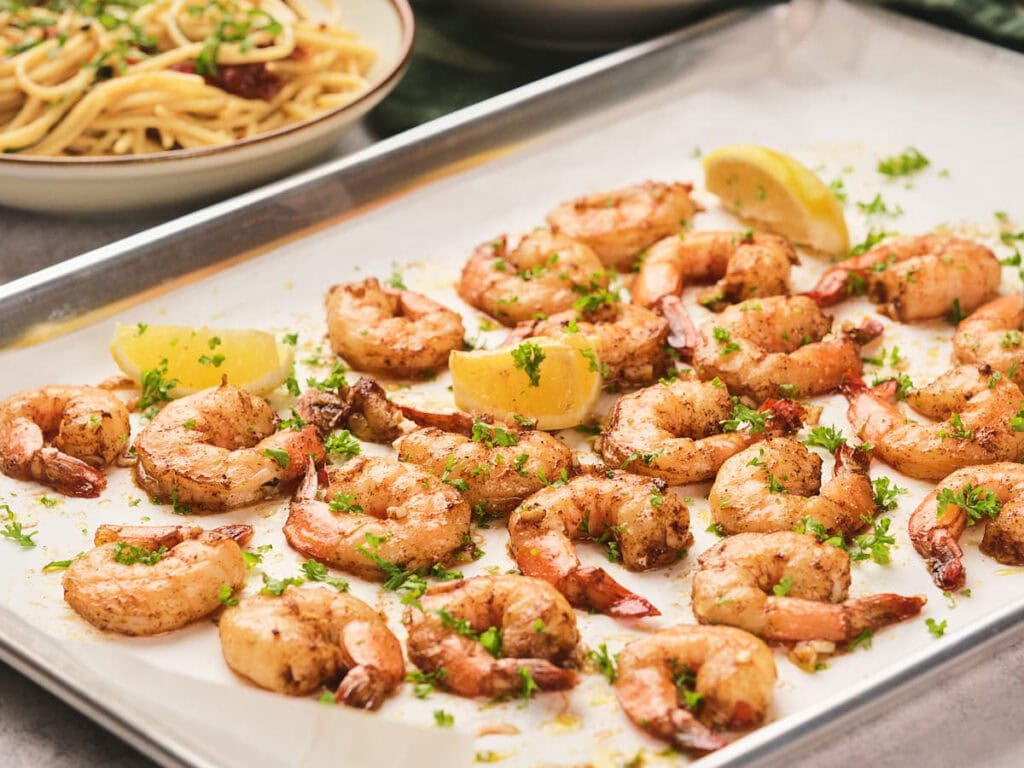 A tray of seasoned shrimp garnished with herbs and fresh lemon wedges, next to a plate of pasta.
