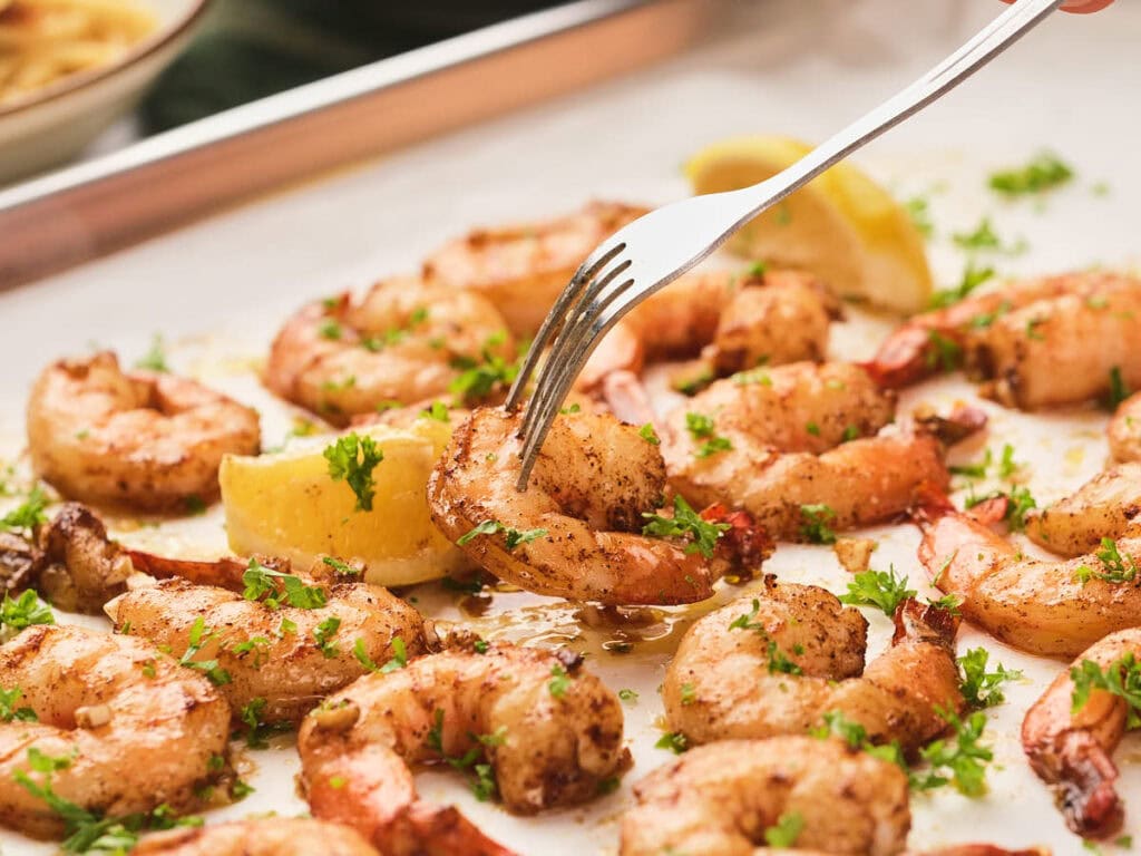 A close-up shot shows a fork picking up a seasoned shrimp from a tray, garnished with fresh herbs and lemon wedges.