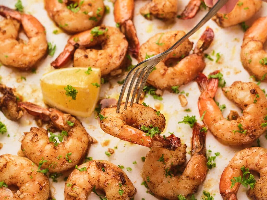 A tray of baked shrimp garnished with chopped herbs and lemon wedges is shown with one shrimp being picked up by a fork.