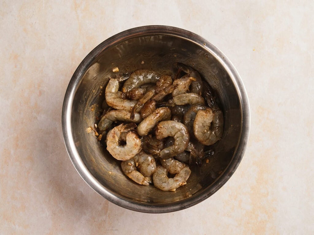 Raw shrimp marinated with spices and garlic in a stainless steel mixing bowl.