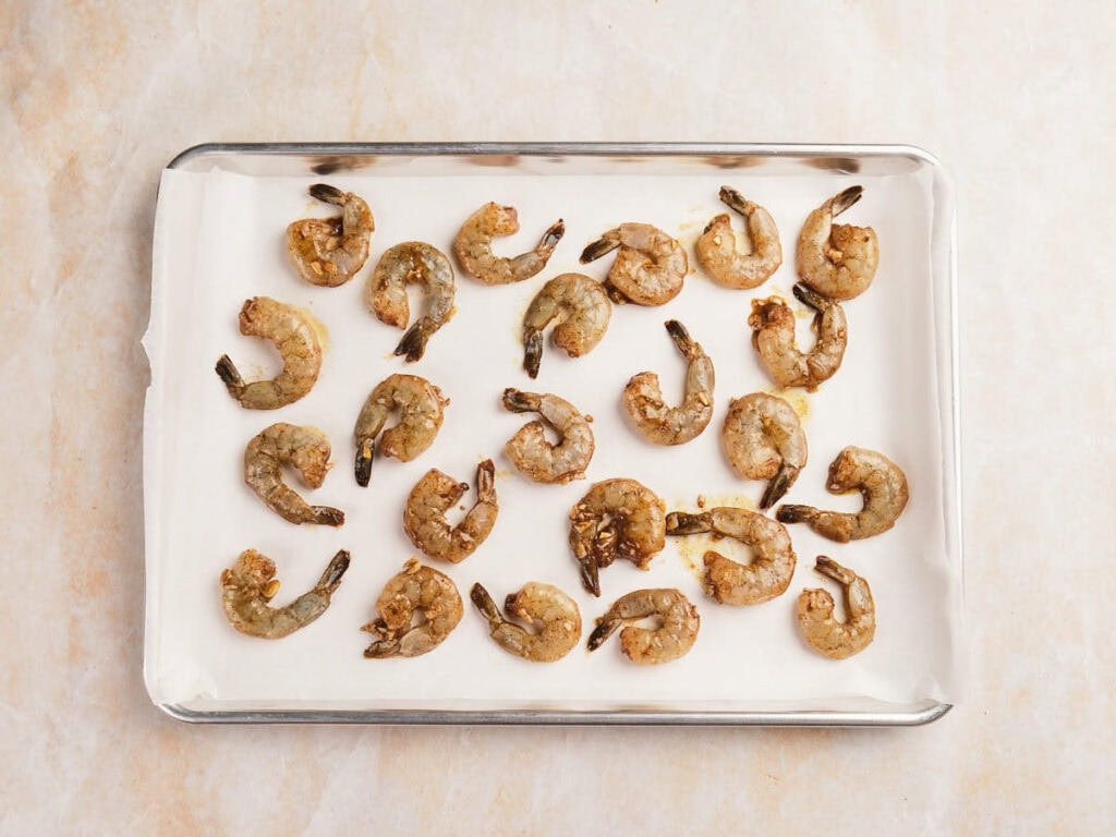 Raw marinated shrimp arranged in rows on a parchment-lined baking sheet on a light surface.
