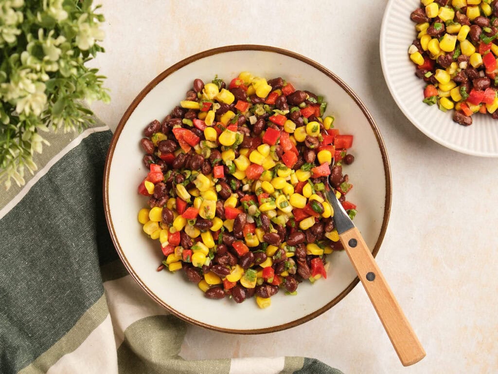 A bowl of black bean salad with a fork, next to a striped napkin and a smaller plate of the same salad.